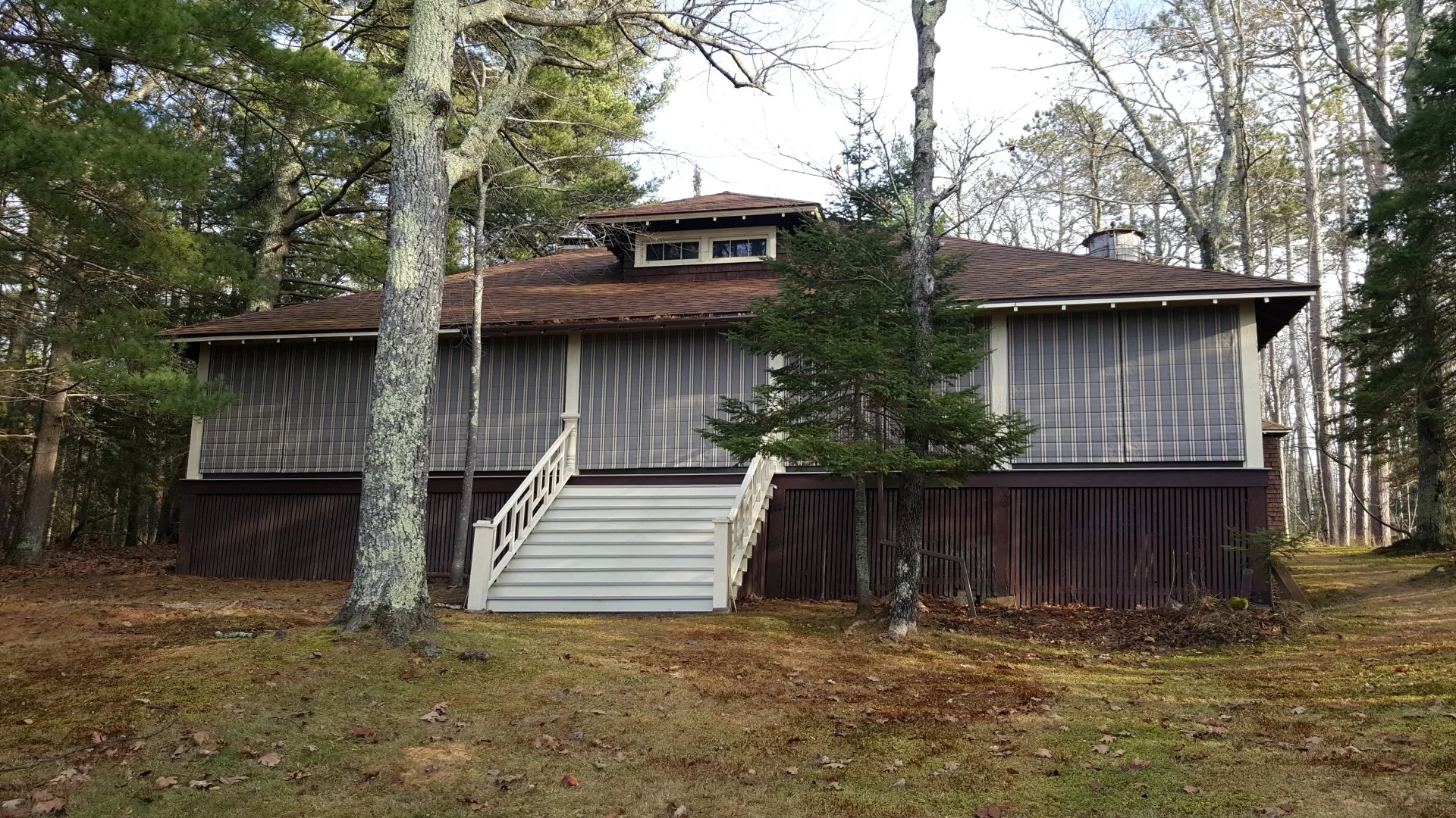 A wooden house with a large screened porch, elevated stairs, and Weather Queen Shades for added comfort, nestled among trees in a forested area.