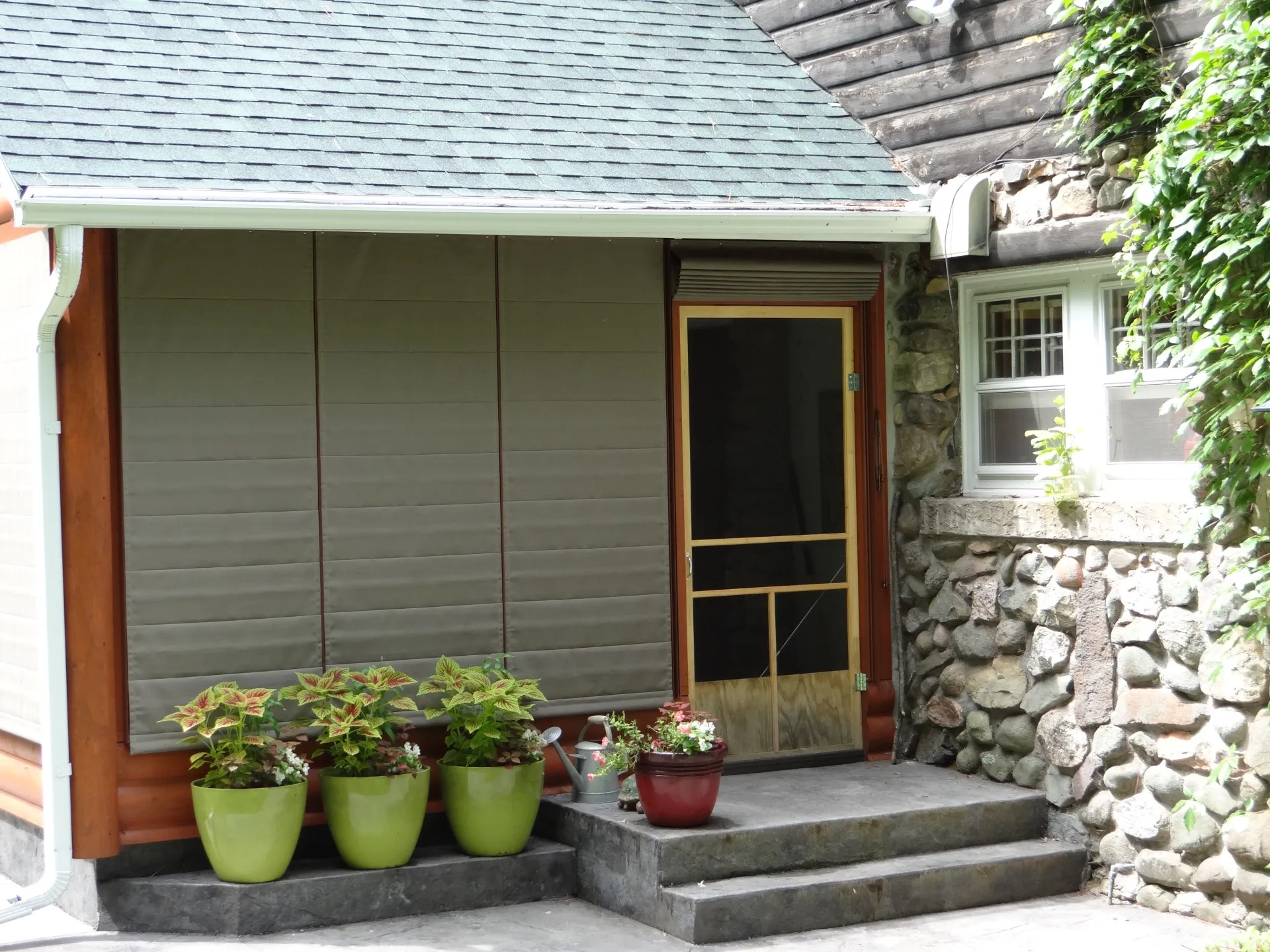 Have to love the small casement windows and vine covered walls. (3) A stone and wood house exterior with a screened porch door, four potted plants on the front steps, and windows partially covered by Weather Queen Shades.