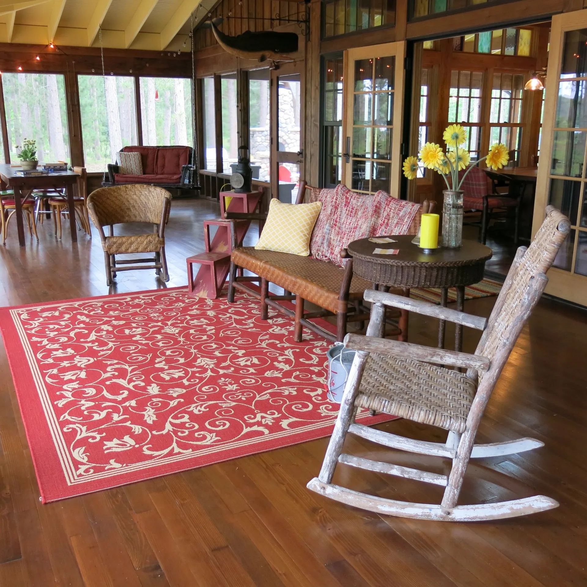 Sunny porch room with wooden floors, wicker furniture, a red patterned rug, a rustic rocking chair, and a table with yellow flowers. Large windows are lined with Weather Queen Shades to let in just the right amount of light.