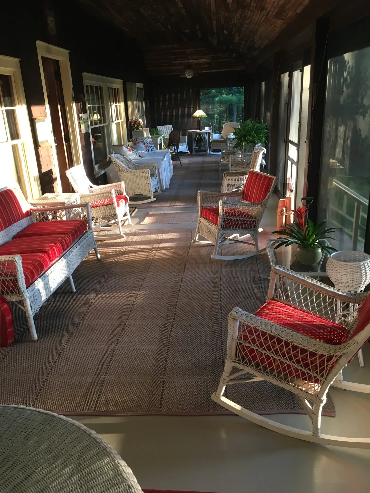 IMG_1521 (1) (2) A long, screened porch with wicker furniture featuring red cushions, a rug, potted plants, and sunlight streaming through windows filtered by elegant Weather Queen Shades.