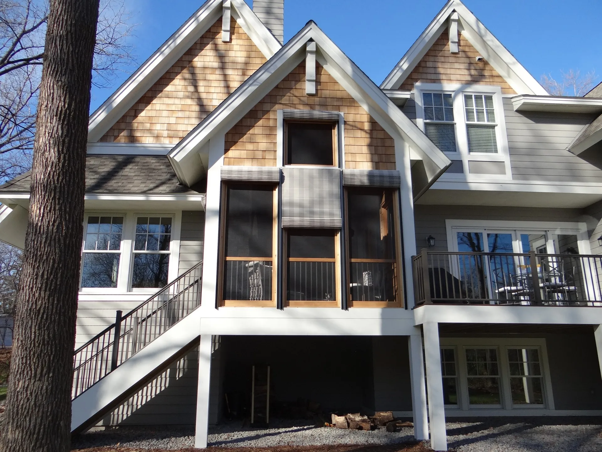 Two-story house with wood siding, screened porch featuring Weather Queen Shades, exterior stairs to an elevated entrance, and a ground-level covered area with firewood stored underneath.