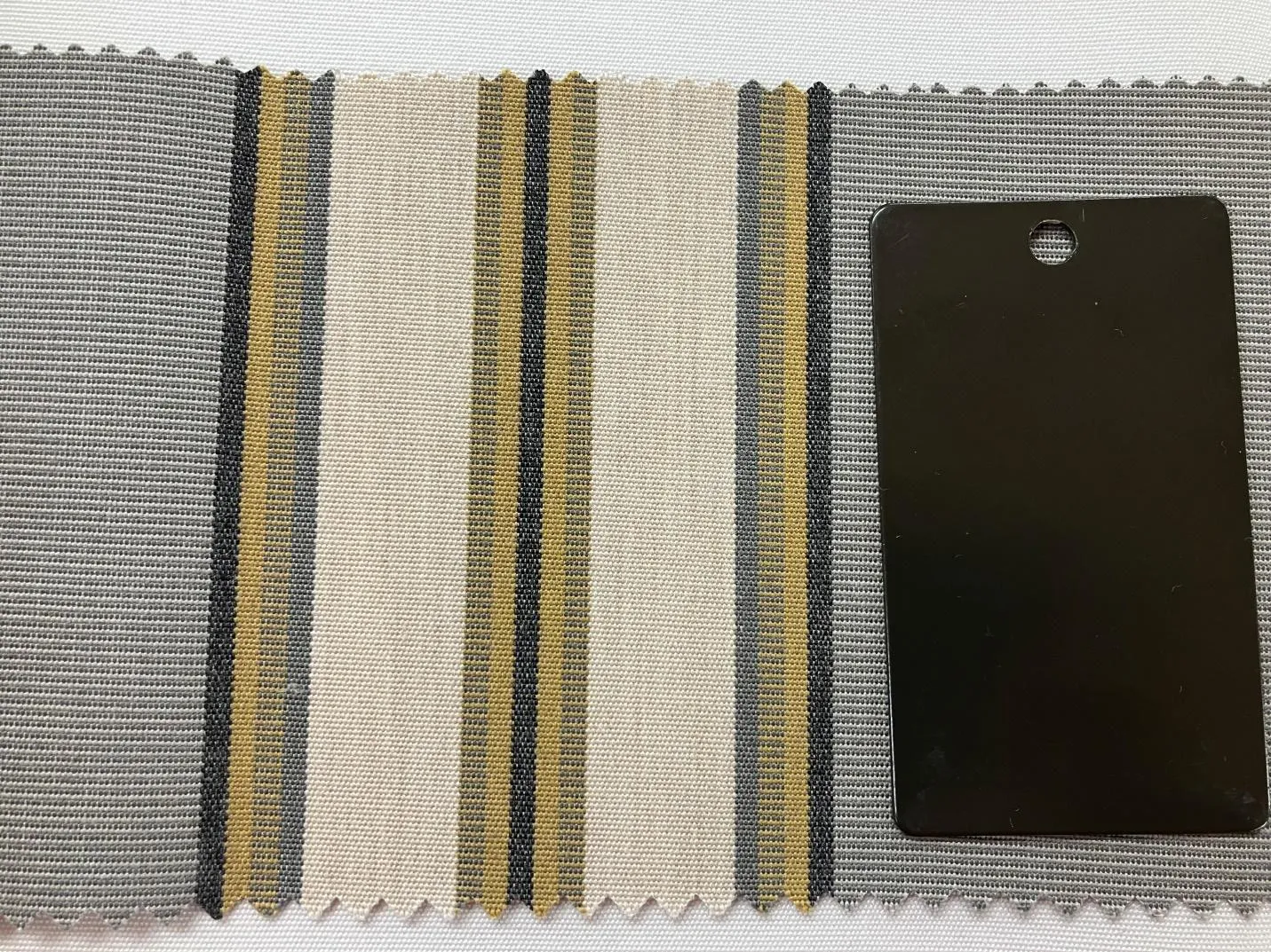 image5 A fabric swatch with wide vertical stripes in beige, gray, yellow, and black evokes the tranquil tones of Lake Superior near Madeline Island, next to a solid black rectangular sample with a hole near the top edge.
