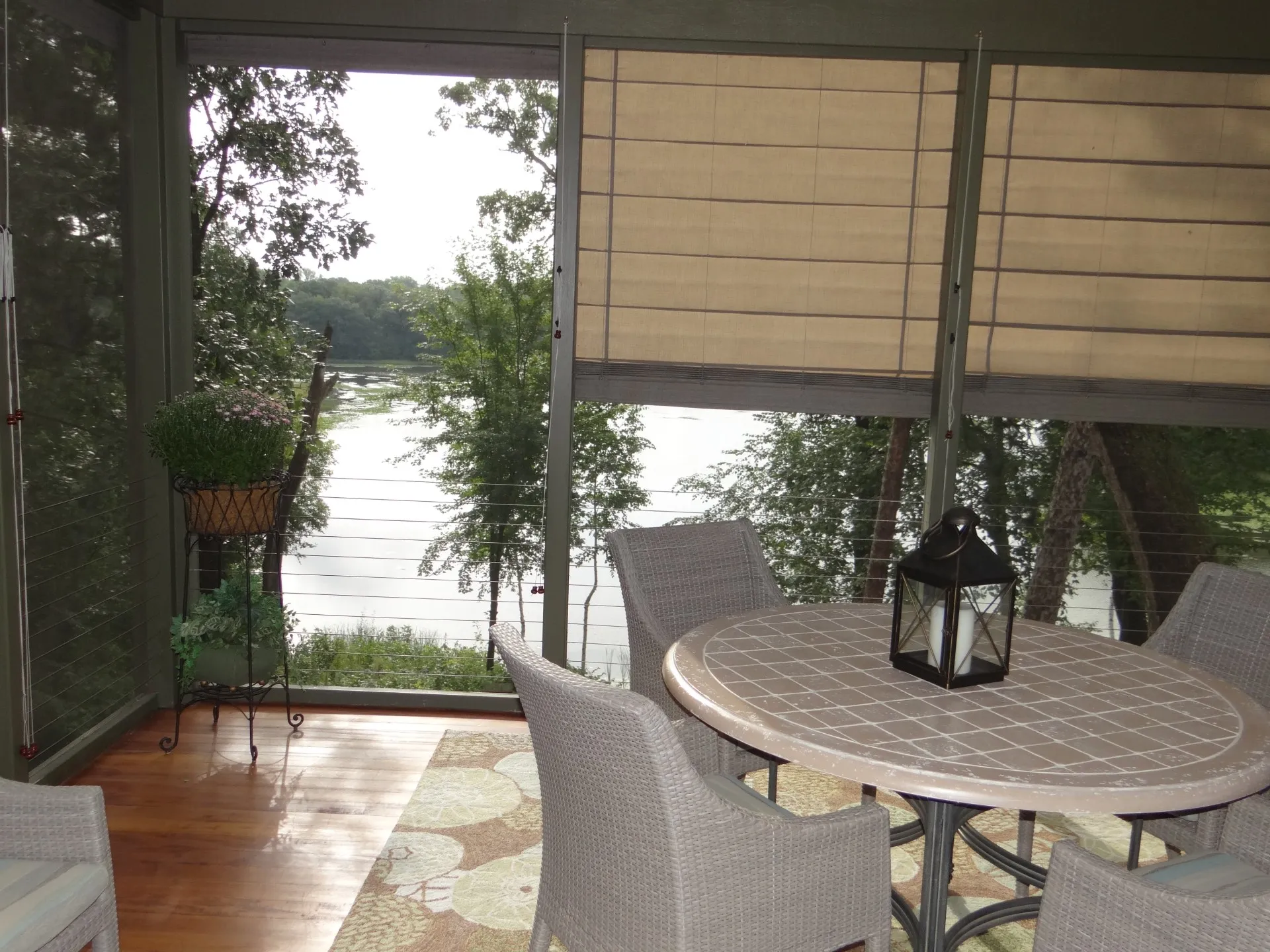 Fully decorated their porch and ready for morning coffee A sunlit screened porch with a round table, four chairs, a black lantern, potted plants, and woven shades partially rolled down, overlooking trees and a lake in the background.