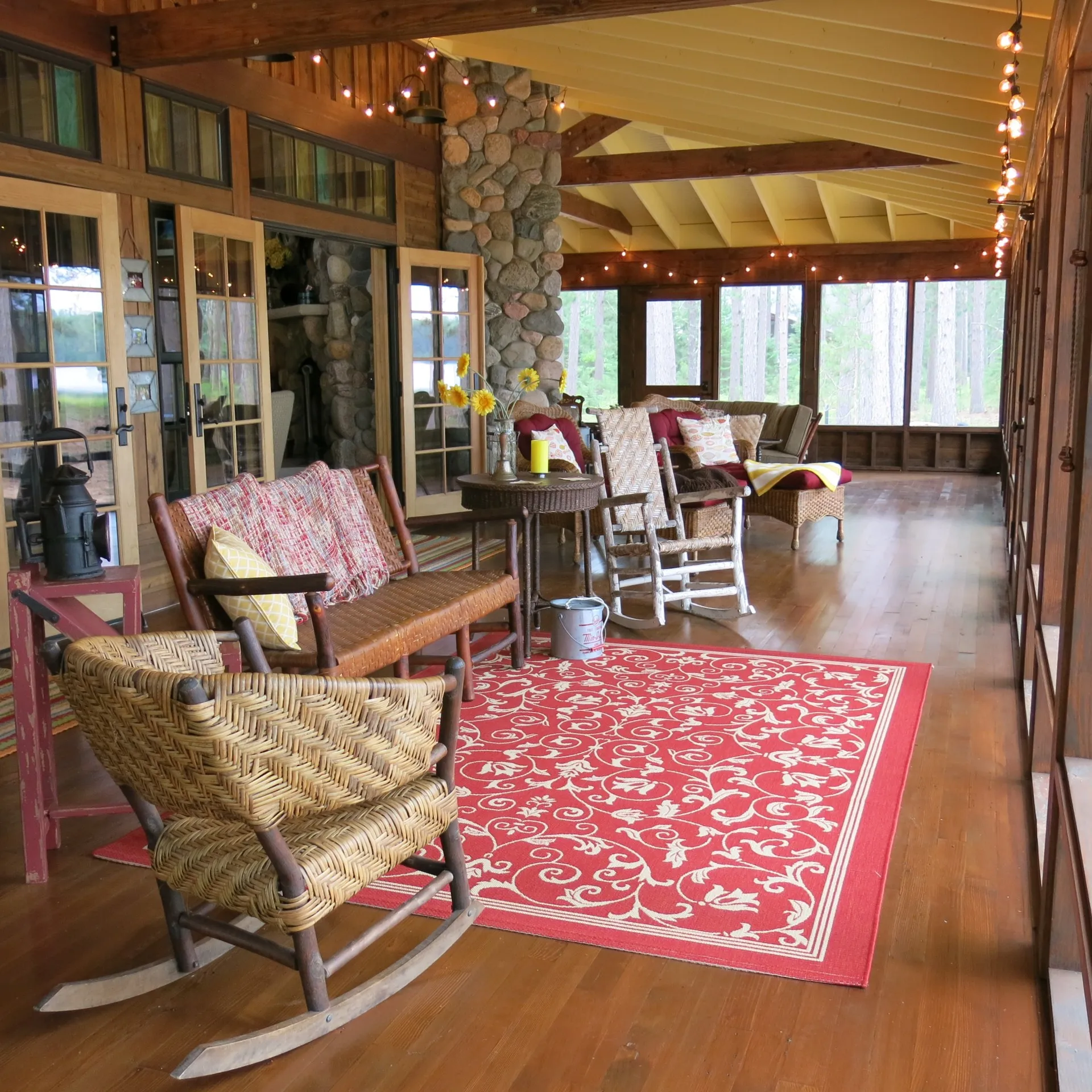 Reminiscent of a simpler time. A cozy screened porch with wooden floors, wicker rocking chairs, a red patterned rug, cushioned seating, a stone fireplace, string lights, and large windows overlooking trees.