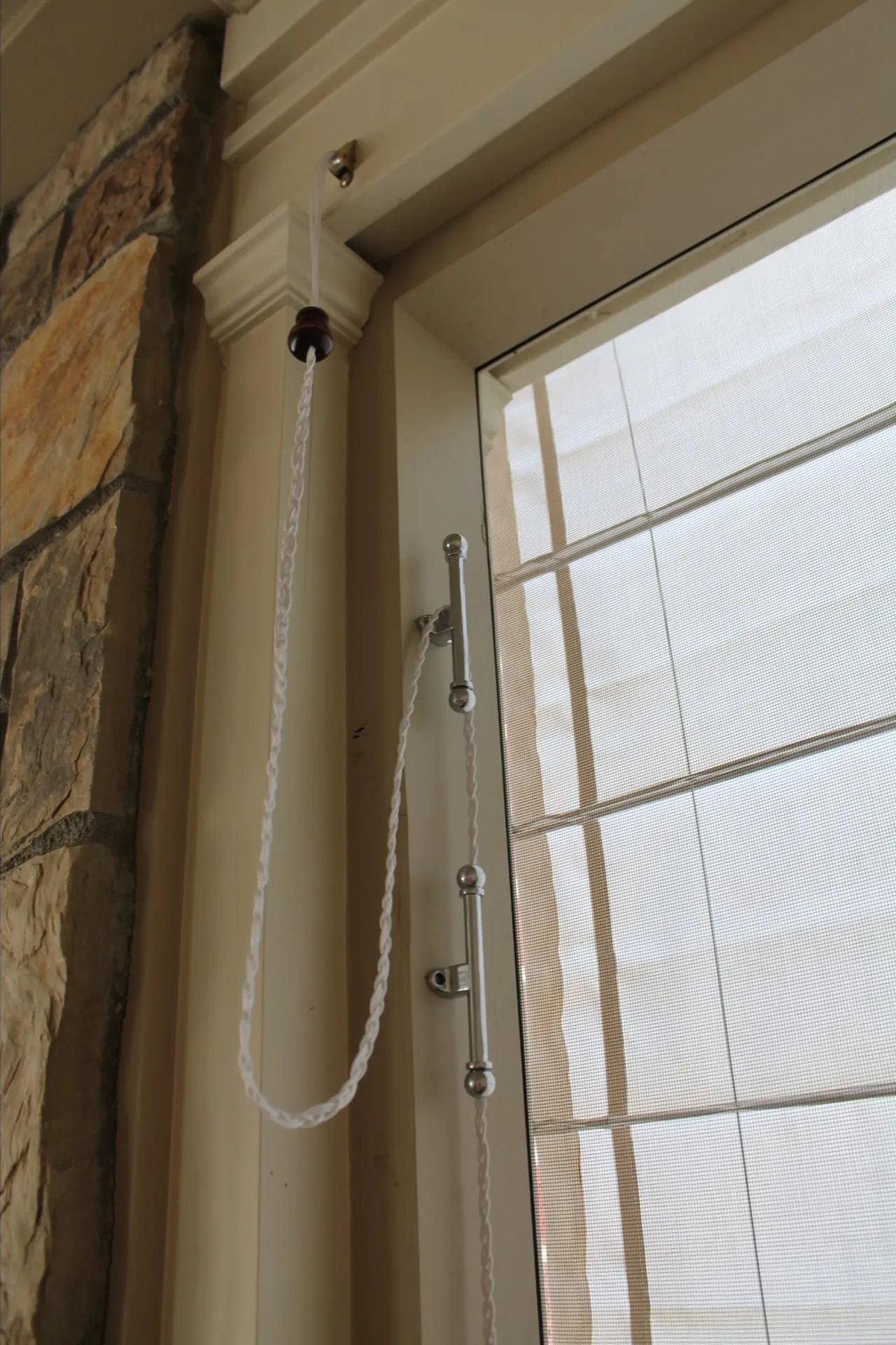 A close-up of a window with a Roman shade, showing a white cord looped around a metal cleat attached to the window frame. Part of a stone wall is visible on the left side.