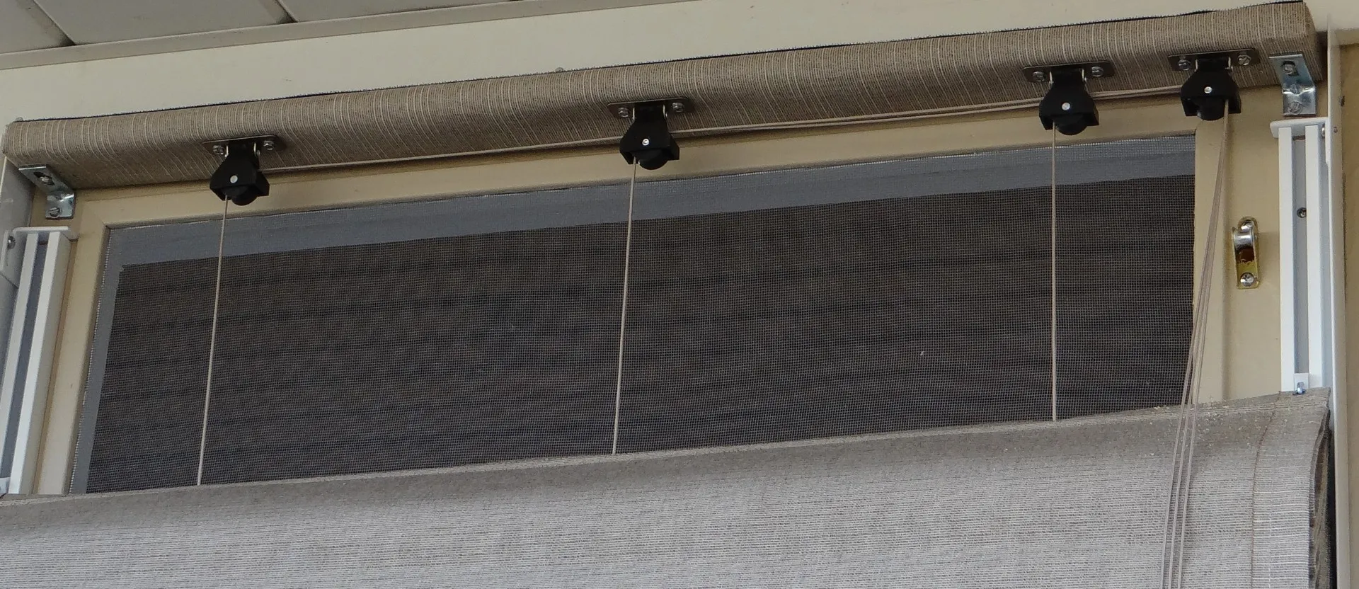 Close-up of a window with a rolled-up beige fabric blind, metal brackets, pulleys, and cords for adjusting the blind’s height. The window frame and a mesh screen are also visible.